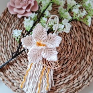 Könnte beinhalten: Eine weiße Makramee-Blume mit gelbem Zentrum und einem langen Fransen. Die Blume hängt an einem Zweig mit weißen Blüten.