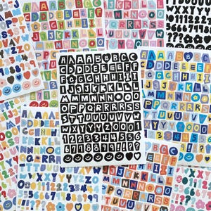Könnte beinhalten: Eine Sammlung bunter Buchstaben- und Zahlenaufkleber in verschiedenen Größen und Schriftarten. Die Aufkleber zeigen Buchstaben, Zahlen und Symbole in einer Vielzahl von Farben, darunter Schwarz, Weiß, Rot, Blau und Gelb. Die Aufkleber sind auf Bögen angeordnet.