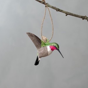 May include: A green and grey hummingbird ornament with a red throat hangs from a twig by a twine string.
