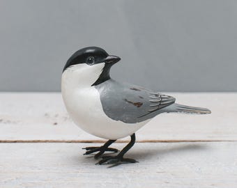 May include: A small, gray and white bird figurine with black eyes and a black head. The bird is standing on a white wooden surface.
