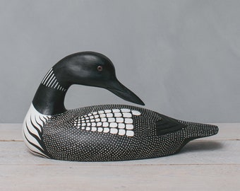 May include: A hand-carved wooden loon figurine. The bird features a black head and back, with white accents on the neck and body. The body is detailed with small white dots. The loon has a long, black beak and a red eye.