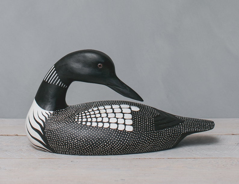 May include: A black and white hand-carved wooden loon figurine. The loon is in a resting position with its head turned to the side. The figurine is detailed with black and white paint and has a realistic appearance.