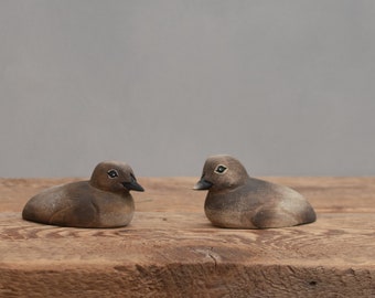 May include: Two small, hand-carved wooden birds, painted in shades of brown and gray, are facing each other on a rustic wooden surface.