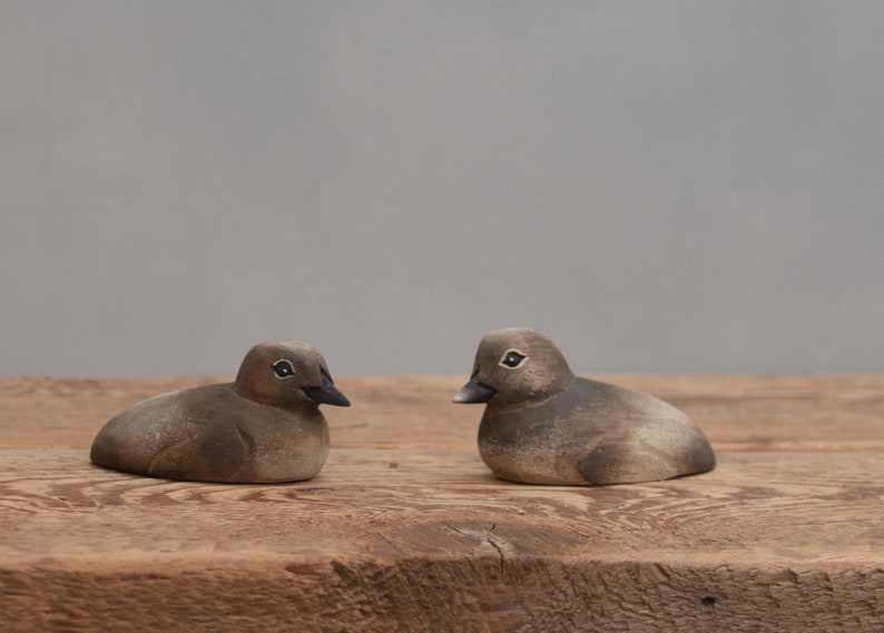Puede incluir: Dos peque&ntilde;os p&aacute;jaros de madera tallados a mano, pintados en tonos marrones y grises, se miran en una superficie de madera r&uacute;stica.