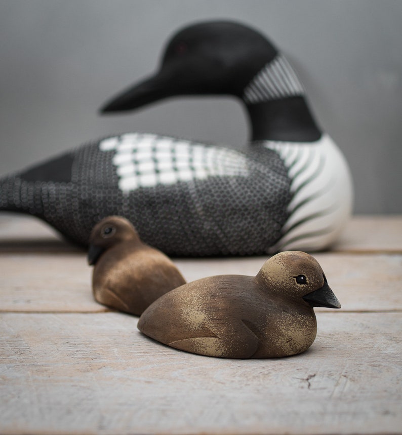 May include: Three wooden bird carvings, a black and white loon and two brown ducks. The loon is larger than the ducks and is positioned behind them. The ducks are facing the camera.