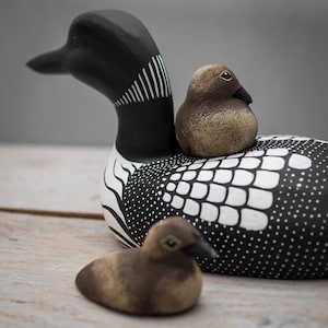 May include: A black and white painted wooden loon with a brown and white painted bird perched on its back. A smaller brown and white painted bird is sitting on the table in front of the loon.