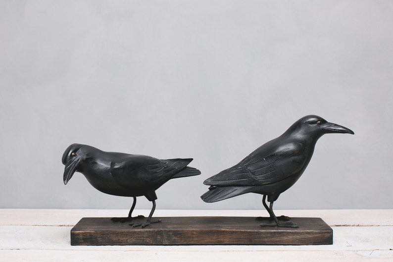May include: Two black crow sculptures perched on a wooden base. The crows are facing opposite directions, one looking to the left and the other to the right.