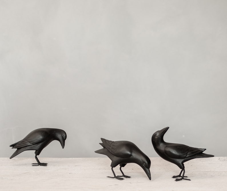 May include: Three black crow figurines, each with a different pose, are arranged on a white surface. The crows are made of a smooth, dark material and have realistic details.