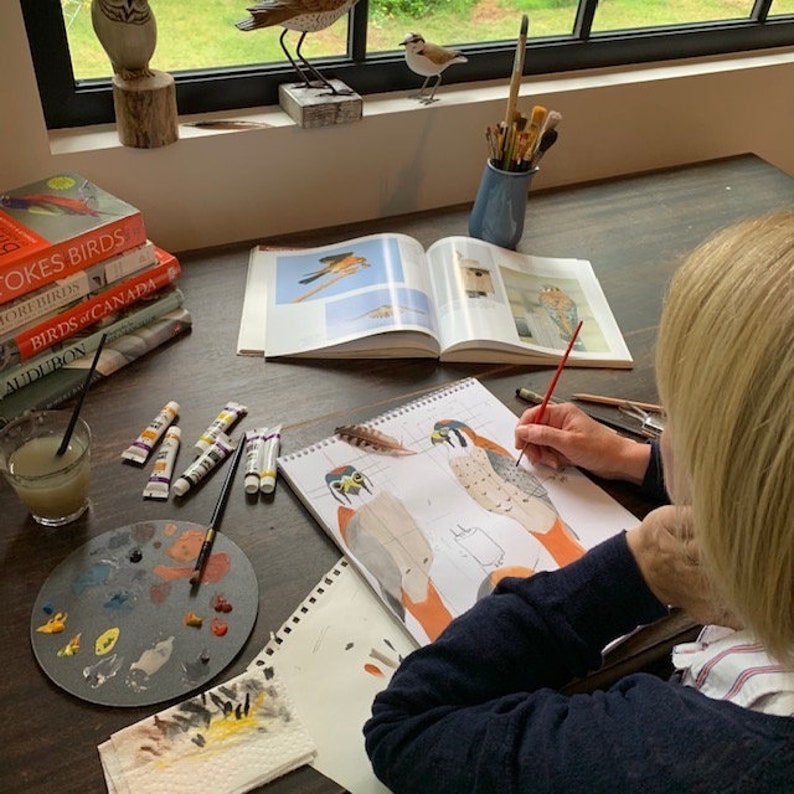 May include: A person is painting a detailed illustration of two birds in a sketchbook. The sketchbook is open on a wooden table with a glass of juice, paint tubes, a paint palette, and a stack of bird books. The books are titled "Stokes Birds", "More Birds", "Birds of Canada", and "Audubon".
