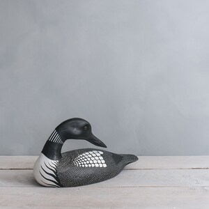 May include: A black and white wooden loon figurine with a detailed design. The figurine is lying on a white wooden surface.
