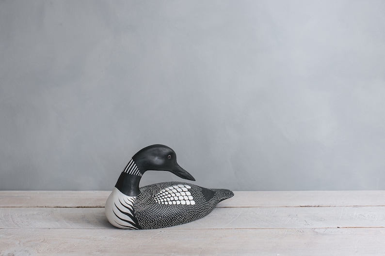 May include: A black and white wooden loon figurine with a detailed design. The figurine is lying on a white wooden surface.