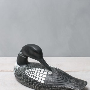 May include: A black and white wooden loon figurine with a spotted pattern and a white grid pattern on its back. The loon is in a resting position with its head tucked down.