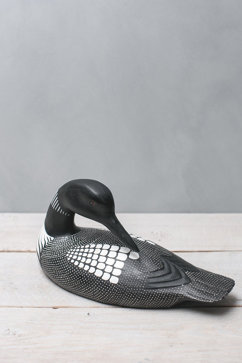 May include: A black and white wooden loon figurine with a spotted pattern and a white grid pattern on its back. The loon is in a resting position with its head tucked down.