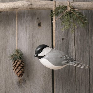 May include: A carved wooden chickadee ornament with a black head, gray wings, and a white chest, hanging from a jute string attached to a branch. A small pinecone and a sprig of evergreen needles are also suspended from the same branch.