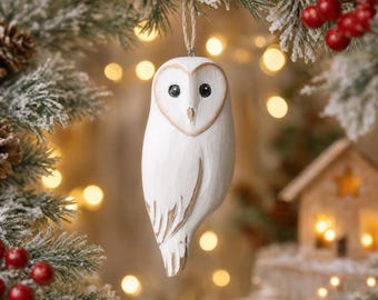 Puede incluir: Un adorno de madera blanca con forma de lechuza, con ojos negros y pico puntiagudo, que presenta detalles de plumas talladas con acentos marrones. La lechuza está suspendida por un fino lazo de cuerda de yute para colgar.