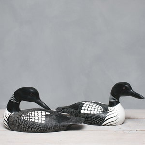 Puede incluir: Dos tallas de madera de p&aacute;jaro loon en blanco y negro. El loon de la izquierda est&aacute; mirando hacia la derecha y el loon de la derecha est&aacute; mirando hacia la izquierda. Ambos loons tienen ojos negros y marcas blancas.