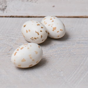 May include: Three speckled white eggs with brown spots on a white wooden surface.
