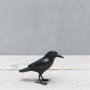 May include: A black crow figurine on a white wooden surface. The crow is facing to the right and has a detailed beak and feet.