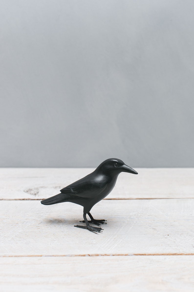 May include: A black crow figurine on a white wooden surface. The crow is facing to the right and has a detailed beak and feet.