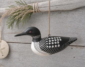 May include: A hand-painted wooden loon ornament featuring a black head with a red eye, a white chest with black stripes, and a black body with white square and dot patterns. The bird hangs from a brown twine string attached to a metal eyelet.