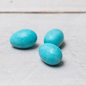 May include: Three turquoise oval sweets are arranged on a light-coloured, textured wooden surface. The sweets have a smooth, slightly glossy appearance. The image is well-lit, highlighting the vibrant colour of the sweets.