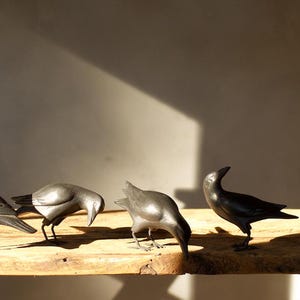 May include: Four black crow figurines are arranged on a wooden surface. The crows are in various poses, with one looking up, one looking down, and two looking straight ahead.