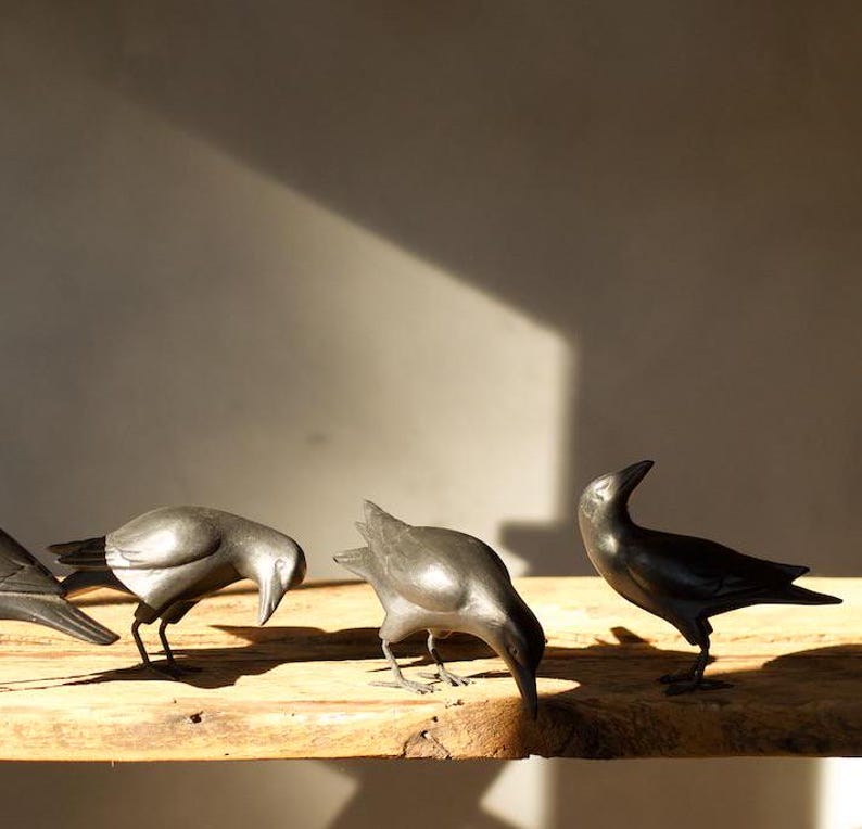 May include: Four black crow figurines are arranged on a wooden surface. The crows are in various poses, with one looking up, one looking down, and two looking straight ahead.