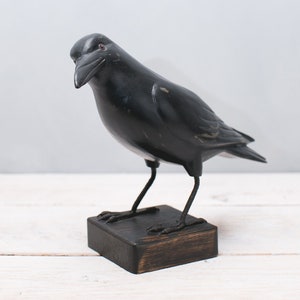 May include: Black wooden crow figurine with a dark brown wooden base. The crow is perched on the base with its wings spread out.