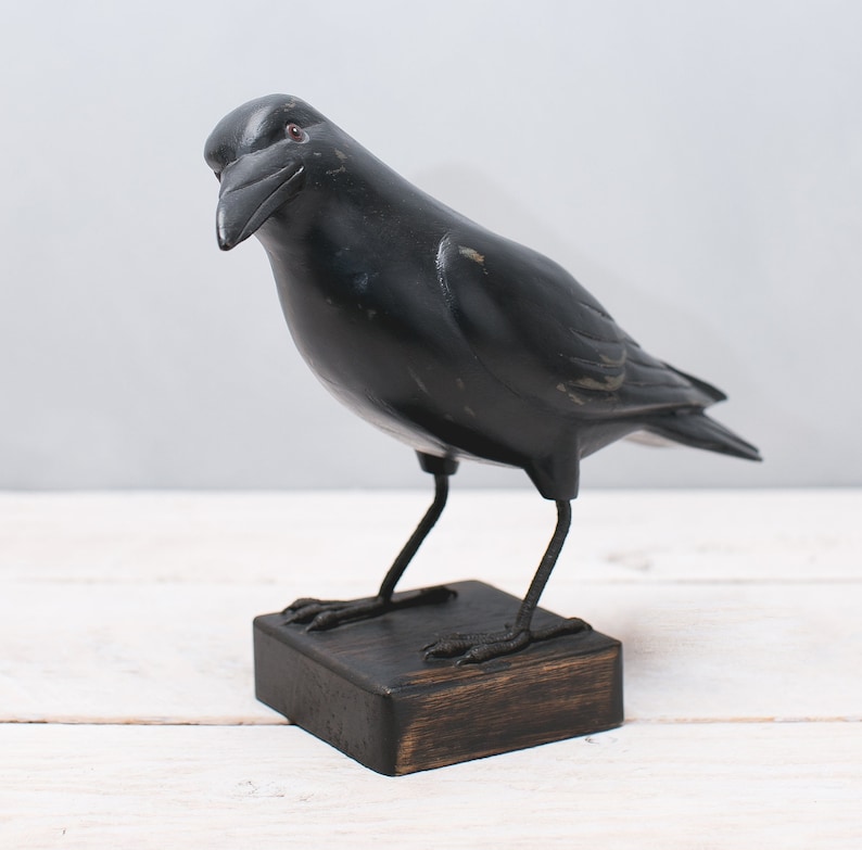 May include: Black wooden crow figurine with a dark brown wooden base. The crow is perched on the base with its wings spread out.