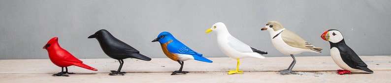 May include: A collection of seven hand-painted wooden bird figurines. The birds are painted in various colors, including red, black, blue, white, and brown. The birds are all standing on a light wood surface.