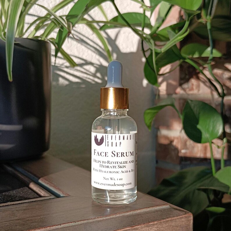 May include: A clear glass bottle of face serum with a blue and gold dropper. The label reads "Face Serum" and lists ingredients. The bottle is 1 fl oz. The background includes green plants and a brick wall.
