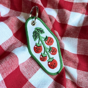 Puede incluir: Un llavero blanco con un borde verde y un diseño de tomates rojos con caras sonrientes en una vid verde. El llavero está sobre un fondo a cuadros rojos y blancos.