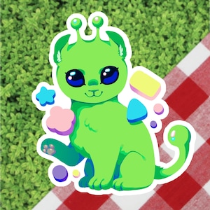 May include: A die-cut sticker featuring a green alien cat with large blue eyes and antennae. The sticker includes colorful star, cloud, and geometric shapes. The background is a red and white checkered pattern.