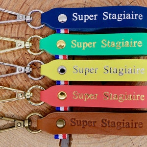 May include: A collection of five leather keychains in various colors: navy blue, green, yellow, coral, and brown. Each keychain has a metal clasp and is embossed with the words "Super Stagiaire" and a star. A small tricolor ribbon is attached.