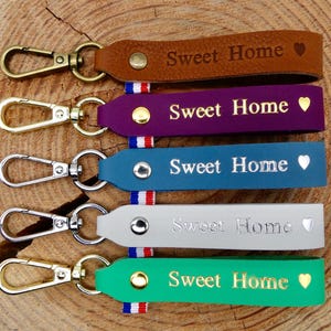 Op de afbeelding: Een verzameling leren sleutelhangers in verschillende kleuren, waaronder bruin, paars, blauwgroen, grijs en groen. Elke sleutelhanger heeft een metalen sluiting en is voorzien van de tekst "Sweet Home" en een hartje.
