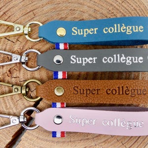 May include: Four leather keyrings in blue, grey, brown, and pink, each with a metal clasp. The keyrings have the text "Super collègue" in gold or silver, with a heart symbol. Each keyring has a small tricolour ribbon.
