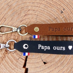 Peut inclure: Deux porte-clés en cuir, un marron et un noir, avec des fermoirs en métal. Le porte-clés marron porte l'inscription "Papa ours" et un symbole de cœur. Le porte-clés noir porte également l'inscription "Papa ours" et un symbole de cœur.
