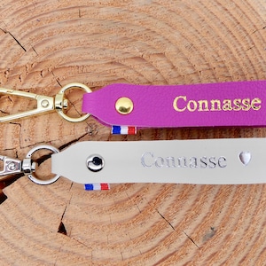 May include: Two leather keychains with metal clasps. One is purple with gold text and a gold heart, the other is white with silver text and a silver heart. Both have a small French flag detail.