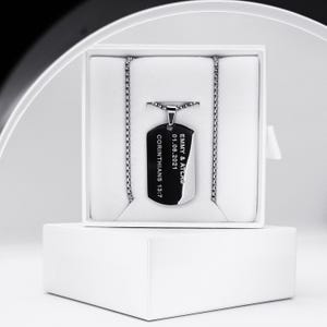 May include: A silver dog tag necklace with a chain, displayed in a white box. The tag is black with the engraved text "EMMY & ATLAS 01.06.2021 CORINTHIANS 13:7". The necklace is presented on a white background.