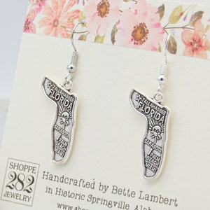 May include: Silver-toned earrings shaped like the state of Florida, with the word "FLORIDA" at the top. The earrings feature a sun design and the names of Florida cities. The earrings are displayed on a card with floral accents.