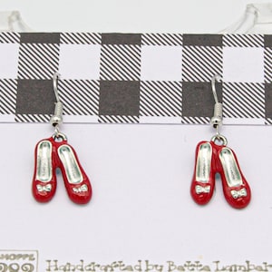 May include: Red enamel dangle earrings shaped like ruby slippers with silver hooks. Each shoe has a small bow and a rhinestone accent. The earrings are displayed on a white card with a black and white checkered pattern.