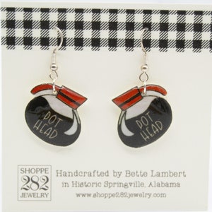 May include: A pair of dangle earrings featuring a black coffee pot design with the words "POT HEAD" in white. The earrings have red and white accents and silver-colored hooks. The earrings are displayed on a white card with a black and white checkered border.
