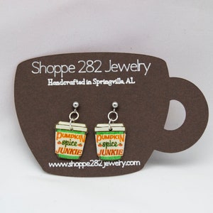May include: A pair of dangle earrings featuring a coffee cup design with the text "Pumpkin Spice Junkie." The earrings are in the shape of a coffee cup with green, orange, and white accents. The earrings are displayed on a brown card.