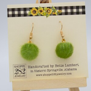 May include: A pair of green pom-pom earrings with gold-colored hooks. The earrings are displayed on a cream-colored card with a black and white checkered border and sunflower accents. The card includes the text "SHOPPE 282 JEWELRY".