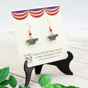 May include: Silver-toned "Marine Mom" earrings with red bead accents. The earrings are displayed on a card with a patriotic red, white, and blue design. The card is held by a black stand, with text that reads "SHOPPE 282 JEWELRY".