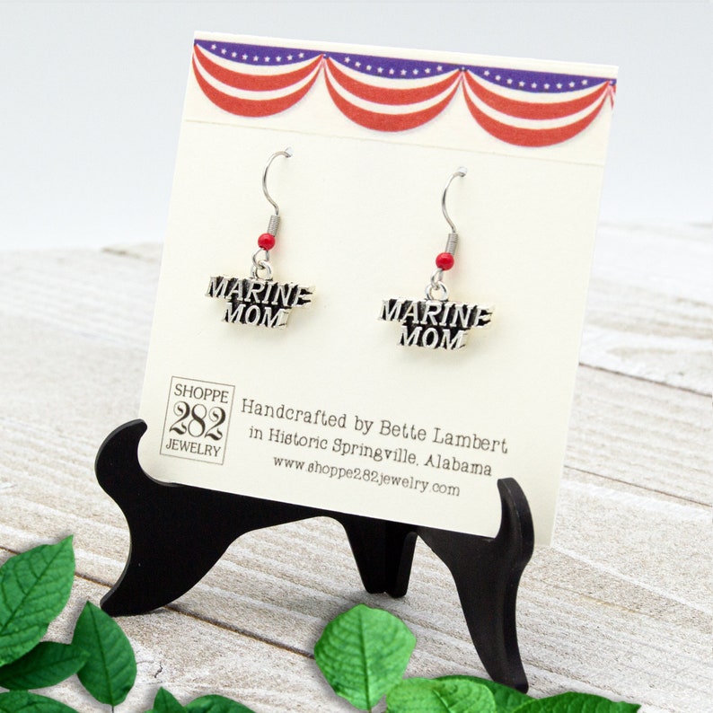 May include: Silver-toned "MARINE MOM" earrings with red bead accents, displayed on a cream-colored card. The card has a patriotic red, white, and blue banner design. The earrings are on a black stand.
