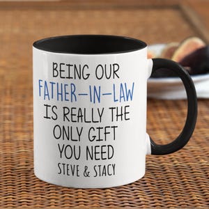 May include: White ceramic mug with a black interior and handle. The mug features the text "BEING OUR FATHER-IN-LAW IS REALLY THE ONLY GIFT YOU NEED STEVE & STACY" in black and blue lettering.
