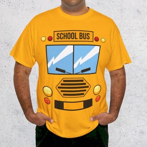 May include: A yellow t-shirt featuring a cartoon school bus graphic on the front. The bus has a blue windscreen, black headlights, and the words "SCHOOL BUS" printed in black above the windscreen.