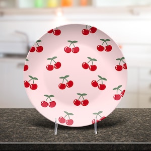 May include: A round, pink ceramic plate with a repeating pattern of red cherries with green stems and leaves. The plate is displayed on a clear stand against a blurred kitchen background.