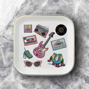 May include: A square white ceramic trinket dish with a gold-toned rim featuring colorful illustrations of 1980s and 1990s items including a pink electric guitar, cassette tapes, a boombox, roller skates, a floppy disk, sunglasses, and a vinyl record.